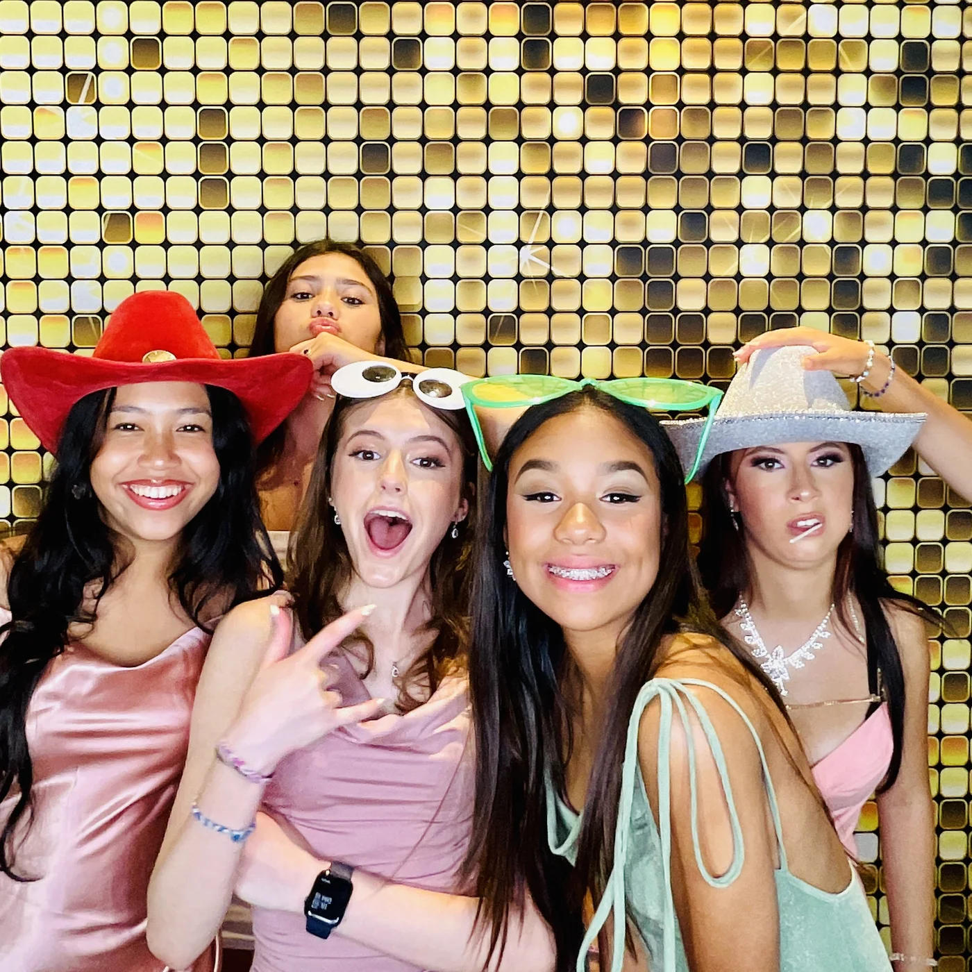 Friends of a quinceañera posing with party props against a gold sequin backdrop